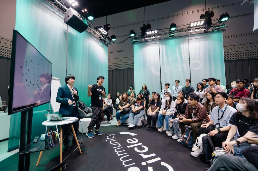 Group of people gathered in a modern event space, seated and standing in a semicircle, listening to two presenters. One presenter gestures while speaking, and the other holds a microphone near a large monitor displaying a network graph. The environment features teal lighting, a black carpet with the words Community Circle, and professional audio-visual equipment overhead.